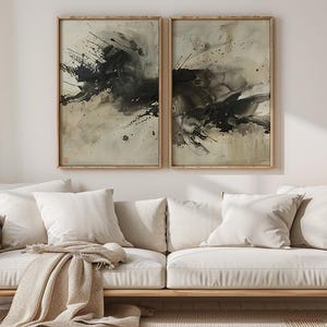 May include: Two framed abstract art prints with black and beige paint splatters hang above a cream-colored sofa with white pillows and a tan throw. A wooden side table holds a vase of dried flowers.