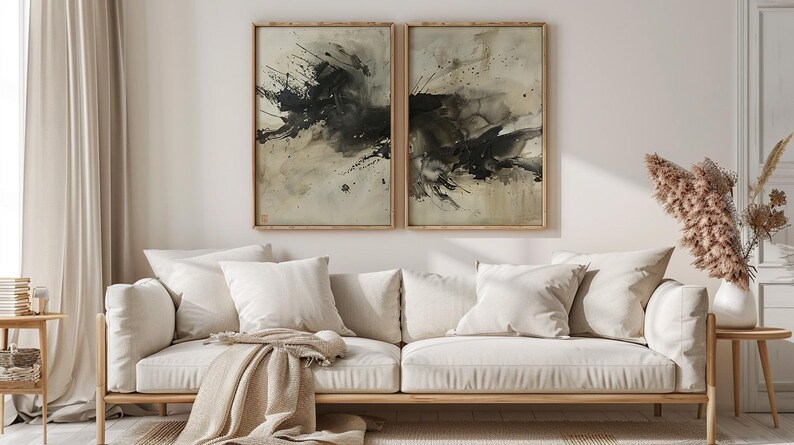 May include: Two framed abstract art prints with black and beige paint splatters hang above a cream-colored sofa with white pillows and a tan throw. A wooden side table holds a vase of dried flowers.