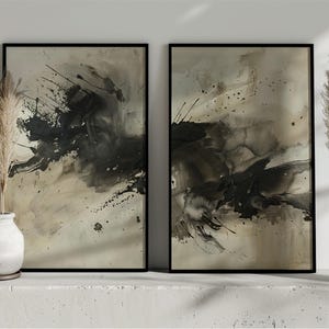 May include: Two framed abstract art prints with black and beige paint splatters. The artwork is displayed on a white surface with decorative pampas grass in white vases on either side. The art has a modern, minimalist aesthetic.