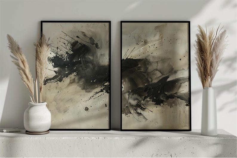May include: Two framed abstract art prints with black and beige paint splatters. The artwork is displayed on a white surface with decorative pampas grass in white vases on either side. The art has a modern, minimalist aesthetic.
