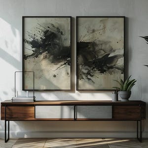 May include: Two framed abstract art pieces with black and gray ink splatters hang above a wooden console table. A potted plant and a decorative metal object sit on the table. A large potted plant with green leaves is on the right.