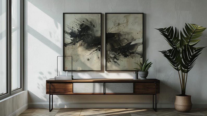 May include: Two framed abstract art pieces with black and gray ink splatters hang above a wooden console table. A potted plant and a decorative metal object sit on the table. A large potted plant with green leaves is on the right.