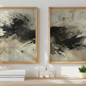 May include: Two framed abstract art prints. Each print features a black ink splatter design on a neutral background. The frames are light wood. The artwork is displayed above a wooden shelf with plants and books.