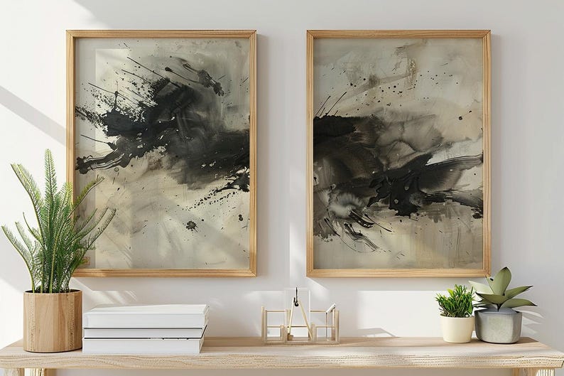 May include: Two framed abstract art prints. Each print features a black ink splatter design on a neutral background. The frames are light wood. The artwork is displayed above a wooden shelf with plants and books.