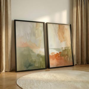May include: Two framed abstract art prints. The artwork features a blend of colours, including green, orange, and beige, creating a landscape-like composition. The frames are black, and the prints are leaning against a wall.