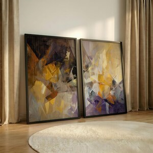 May include: Two framed abstract art prints. The artwork features geometric shapes in shades of gold, purple, and brown. The frames are black. The prints are displayed on a wooden floor with a white rug in front of them.