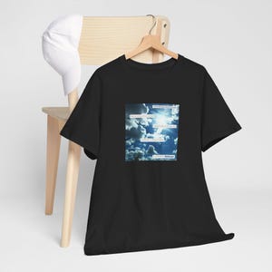 May include: Black t-shirt featuring a square graphic of a blue sky with clouds and text. The text includes phrases such as "Why do I question the moment?" and "Getting to get it over with". A white baseball cap rests on a wooden chair.