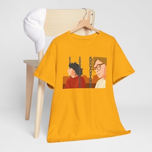 Può includere: T-shirt giallo senape con un'illustrazione di due persone su un'altalena. Una persona indossa un maglione rosso, l'altra occhiali e una camicia bianca. Un berretto da baseball bianco è appoggiato su una sedia di legno.