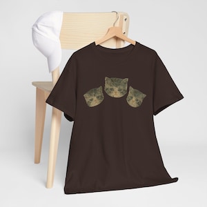 Könnte beinhalten: Dunkelbraunes T-Shirt mit drei Katzengesichtern. Die Katzengesichter haben einen Vintage-Look mit einem Used-Look. Eine weiße Baseballkappe liegt auf einem Holzstuhl im Hintergrund.