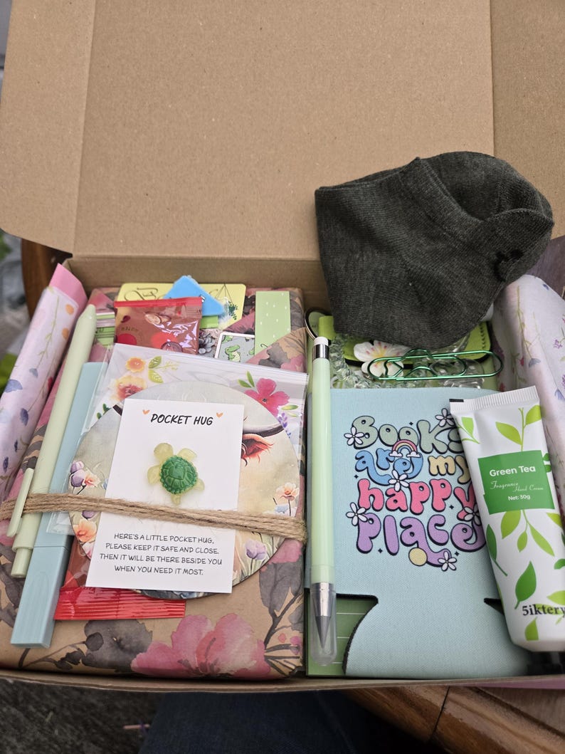 May include: An open cardboard box filled with various items. Includes a green pen, a green and white tube of Green Tea hand cream, a blue drink koozie with the text "Books are my happy place", and a small green turtle pin.