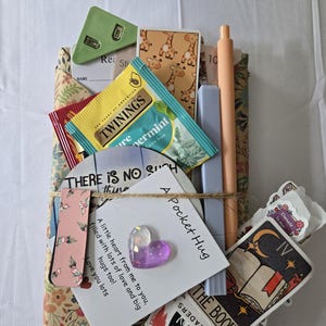 May include: A collection of items wrapped in floral paper, tied with twine. Includes a green clip, tea, stickers, a pen, and a card that says "A Pocket Hug". A purple heart-shaped charm is attached.