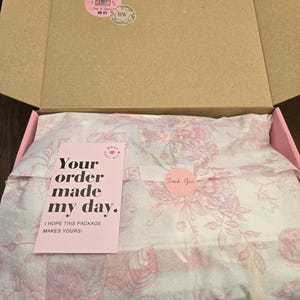 May include: An open cardboard box with a pink interior. Inside, tissue paper with a floral pattern wraps a product. A pink card reads "Your order made my day." A round sticker says "Thank You."