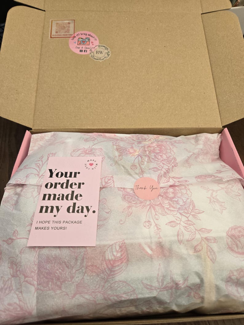 May include: An open cardboard box with a pink interior. Inside, tissue paper with a floral pattern wraps a product. A pink card reads "Your order made my day." A round sticker says "Thank You."