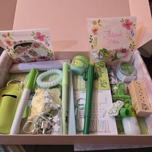 May include: A gift box filled with stationery and self-care items. Includes pens, a hair tie, a keychain, a lip balm, a cactus pen, a stamp, and a card that says "Thank You". The box is pink.