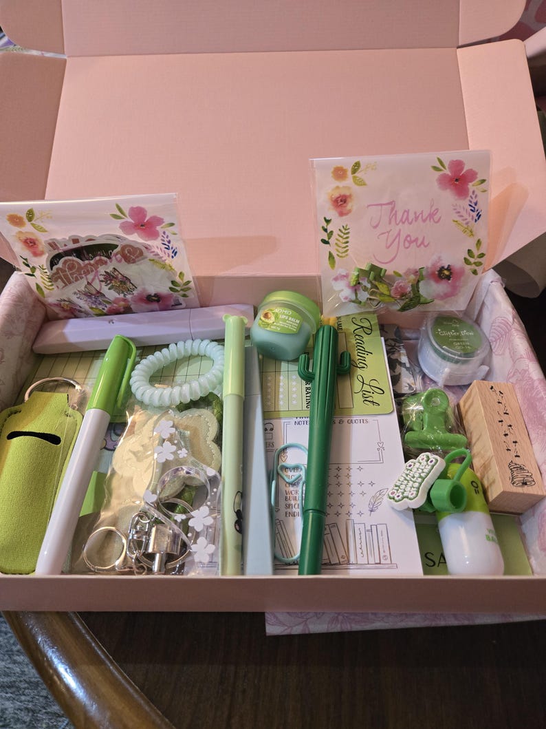 May include: A gift box filled with stationery and self-care items. Includes pens, a hair tie, a keychain, a lip balm, a cactus pen, a stamp, and a card that says "Thank You". The box is pink.
