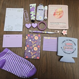 May include: A collection of purple and white stationery and self-care items, including a notepad, pen, lip balm, face mask, and a sock. The items are arranged on a wooden surface, creating a visually appealing assortment.