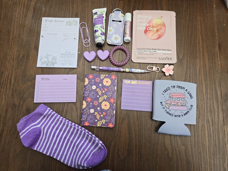 May include: A collection of purple and white stationery and self-care items, including a notepad, pen, lip balm, face mask, and a sock. The items are arranged on a wooden surface, creating a visually appealing assortment.
