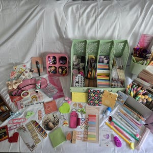May include: A collection of stationery and craft supplies, including pens, stickers, notebooks, and decorative paper. The items are arranged on a white surface, with a variety of colors and patterns visible. There are also storage containers and organizers.
