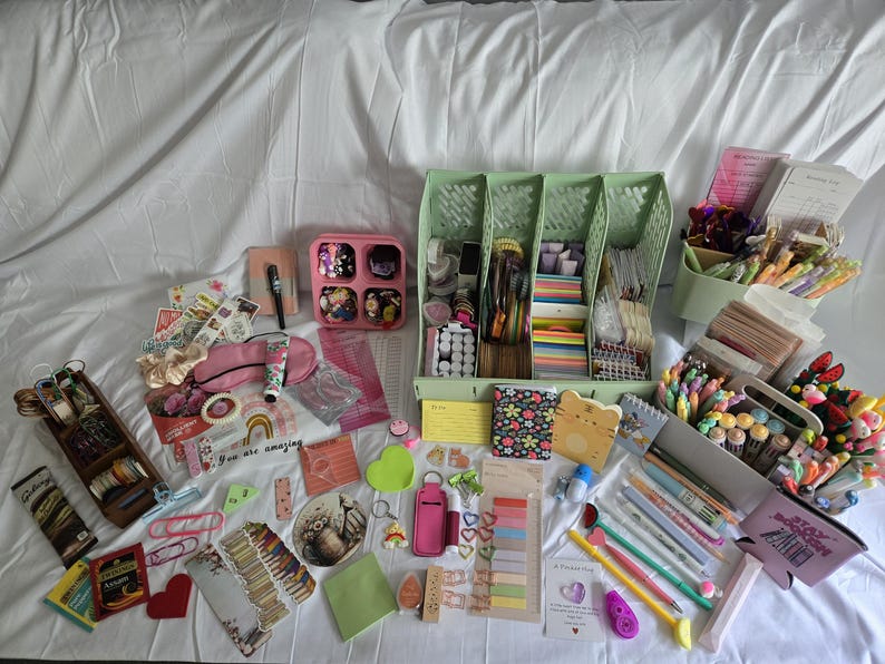 May include: A collection of stationery and craft supplies, including pens, stickers, notebooks, and decorative paper. The items are arranged on a white surface, with a variety of colors and patterns visible. There are also storage containers and organizers.
