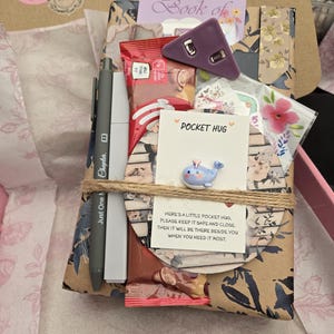 May include: A gift box wrapped in floral paper, tied with twine, containing a pen, snacks, a purple triangle-shaped object, and a card that says "Pocket Hug". The box also includes a small whale figurine.