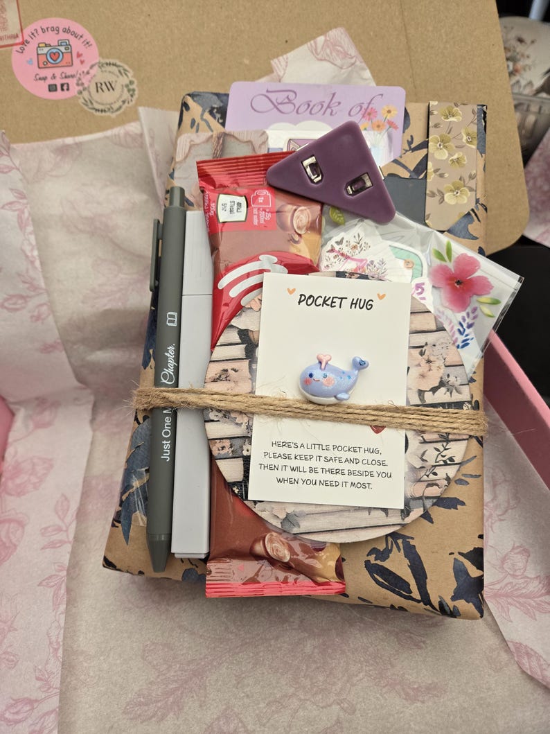 May include: A gift box wrapped in floral paper, tied with twine, containing a pen, snacks, a purple triangle-shaped object, and a card that says "Pocket Hug". The box also includes a small whale figurine.