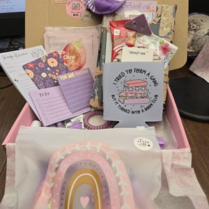 May include: A gift box filled with various items, including a purple and white striped sock, a rainbow-themed pouch with the text "You are amazing", notepads, and a drink holder with the text "I tried to form a gang but it turned into a book club".