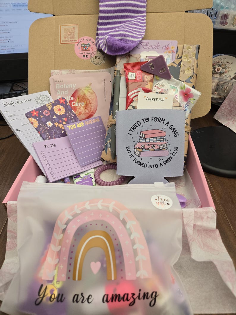 May include: A gift box filled with various items, including a purple and white striped sock, a rainbow-themed pouch with the text "You are amazing", notepads, and a drink holder with the text "I tried to form a gang but it turned into a book club".
