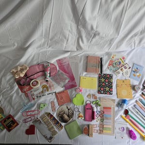 May include: A collection of stationery and small accessories, including notebooks, pens, stickers, and a sleep mask. The items are arranged on a white surface, with a variety of colors and patterns.