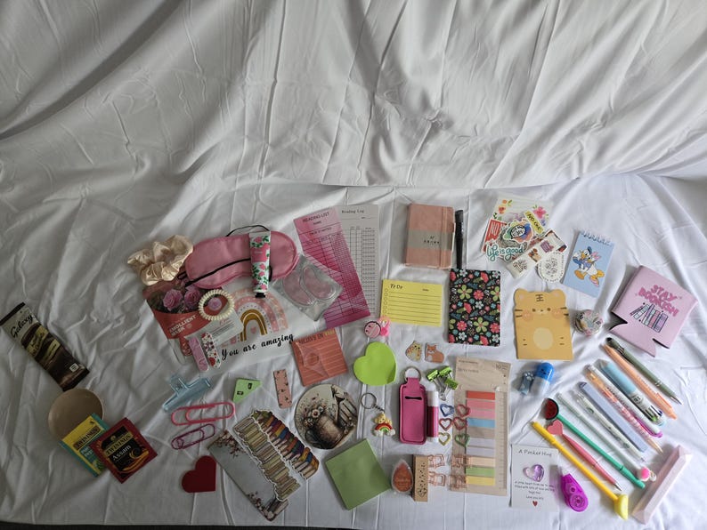 May include: A collection of stationery and small accessories, including notebooks, pens, stickers, and a sleep mask. The items are arranged on a white surface, with a variety of colors and patterns.