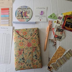 May include: An assortment of stationery and reading accessories, including sticky notes, a reading log, a floral-patterned book cover, and a pen. Also included are stickers, bookmarks, and tea bags. A coaster reads "THERE IS NO SUCH THING AS TOO MANY BOOKS."
