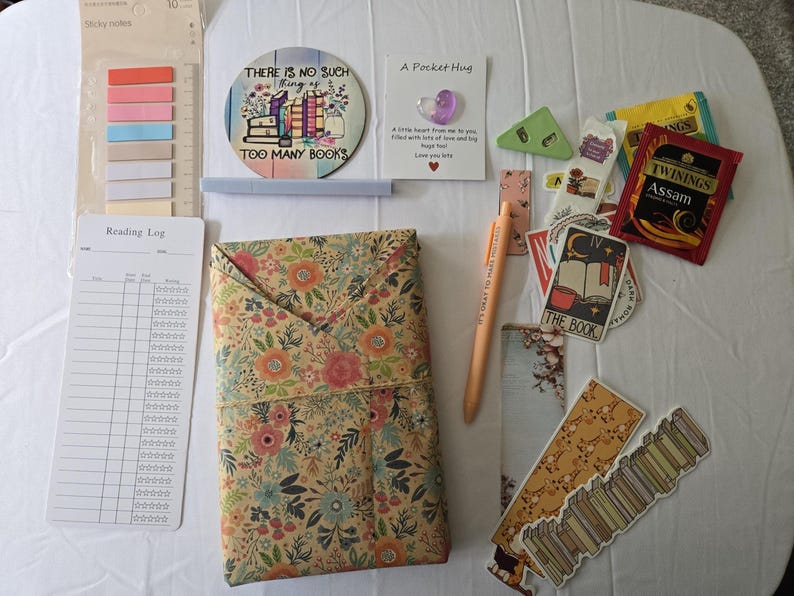 May include: An assortment of stationery and reading accessories, including sticky notes, a reading log, a floral-patterned book cover, and a pen. Also included are stickers, bookmarks, and tea bags. A coaster reads "THERE IS NO SUCH THING AS TOO MANY BOOKS."