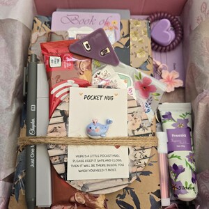 May include: A pink gift box filled with various items, including a pen, a small whale figurine, a purple hair tie, a tube of hand cream, and a card that says "Pocket Hug". The box is tied with twine and contains a variety of patterned papers and small gifts.