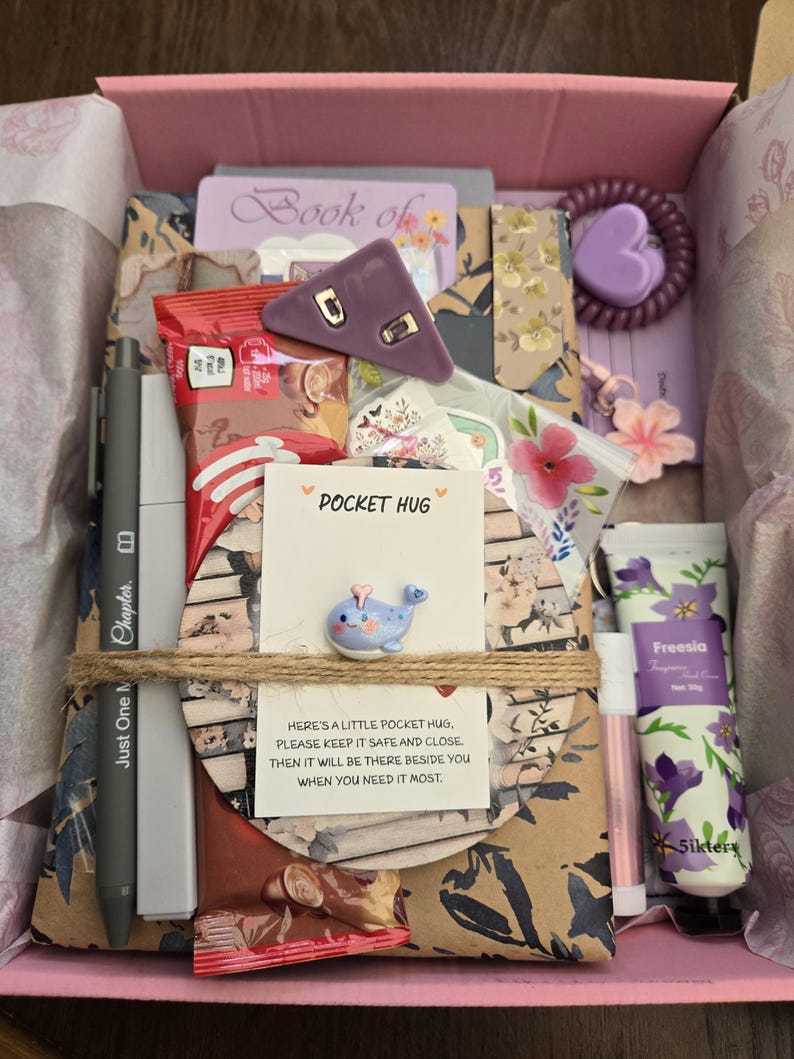 May include: A pink gift box filled with various items, including a pen, a small whale figurine, a purple hair tie, a tube of hand cream, and a card that says "Pocket Hug". The box is tied with twine and contains a variety of patterned papers and small gifts.