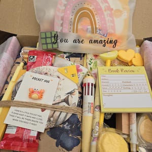 May include: A gift box filled with stationery and small items. Includes a rainbow-shaped decoration, a Rubik's cube, pens, a notepad, and a small crab-shaped pin. The box also contains a card that says "You are amazing."