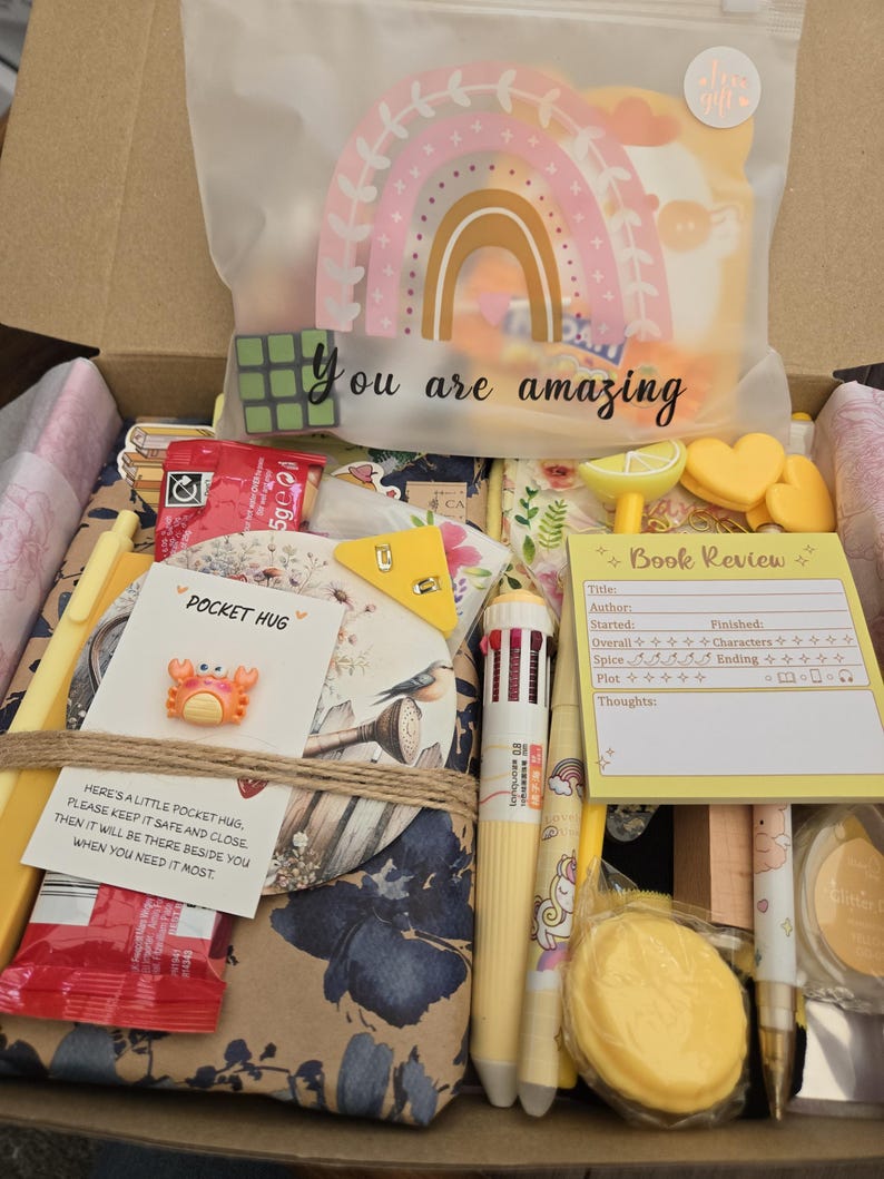 May include: A gift box filled with stationery and small items. Includes a rainbow-shaped decoration, a Rubik's cube, pens, a notepad, and a small crab-shaped pin. The box also contains a card that says "You are amazing."