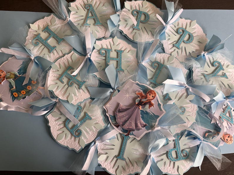 May include: A festive banner with the words "HAPPY BIRTHDAY" spelled out in glittery blue letters. Each letter is attached to a snowflake-shaped cutout with light blue ribbons and tulle accents. Features images of the character Anna from the movie Frozen.