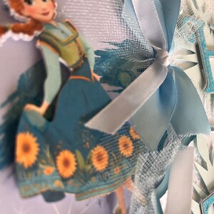 May include: A decorative banner featuring a cartoon image of a character from the movie Frozen. The banner includes light blue ribbons, tulle, and cut-out letters spelling a word. The character is wearing a teal dress with sunflower accents.
