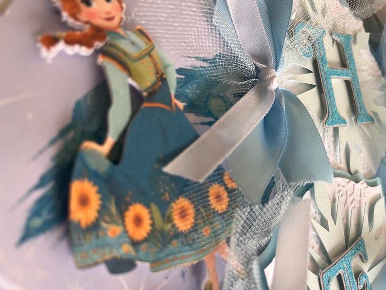 May include: A decorative banner featuring a cartoon image of a character from the movie Frozen. The banner includes light blue ribbons, tulle, and cut-out letters spelling a word. The character is wearing a teal dress with sunflower accents.