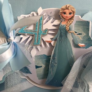May include: A light blue birthday card featuring a cartoon image of Elsa from Frozen. The card has a glittery number 4, white snowflake accents, and light blue ribbons. The overall theme is a Frozen-themed birthday celebration.