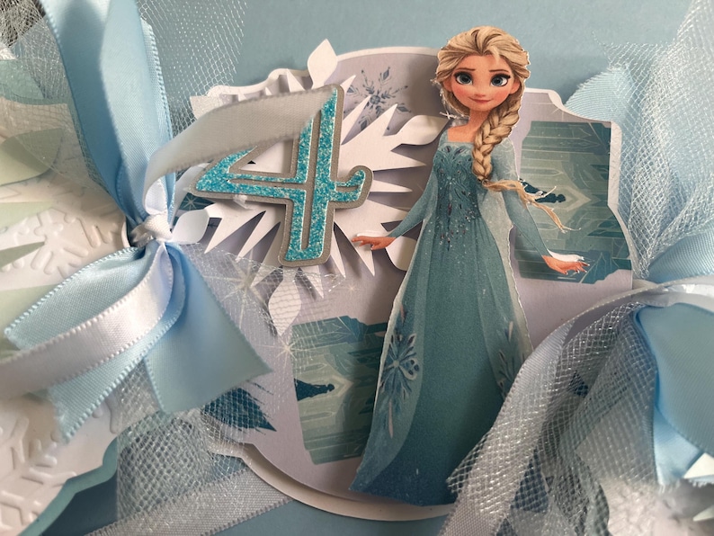 May include: A light blue birthday card featuring a cartoon image of Elsa from Frozen. The card has a glittery number 4, white snowflake accents, and light blue ribbons. The overall theme is a Frozen-themed birthday celebration.