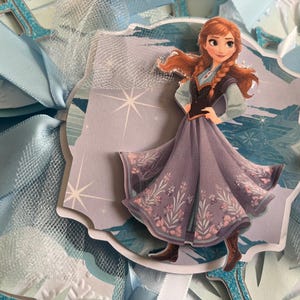May include: A decorative die-cut image of the character Anna from the movie Frozen. Anna is wearing a purple dress with floral accents and brown boots. The image is surrounded by light blue ribbons and tulle.