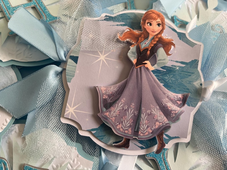 May include: A decorative die-cut image of the character Anna from the movie Frozen. Anna is wearing a purple dress with floral accents and brown boots. The image is surrounded by light blue ribbons and tulle.