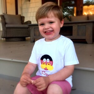 May include: A young child wearing a white t-shirt with a cartoon character design and the name "Cody" printed on it. The child is also wearing pink shorts and is sitting on a porch step.