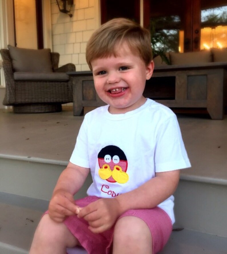 May include: A young child wearing a white t-shirt with a cartoon character design and the name "Cody" printed on it. The child is also wearing pink shorts and is sitting on a porch step.