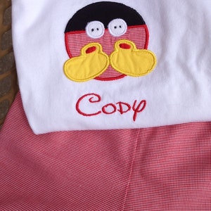 May include: White top with a Mickey Mouse face applique and the name "Cody" embroidered in red. The outfit includes red and white checkered shorts, creating a playful and coordinated look.
