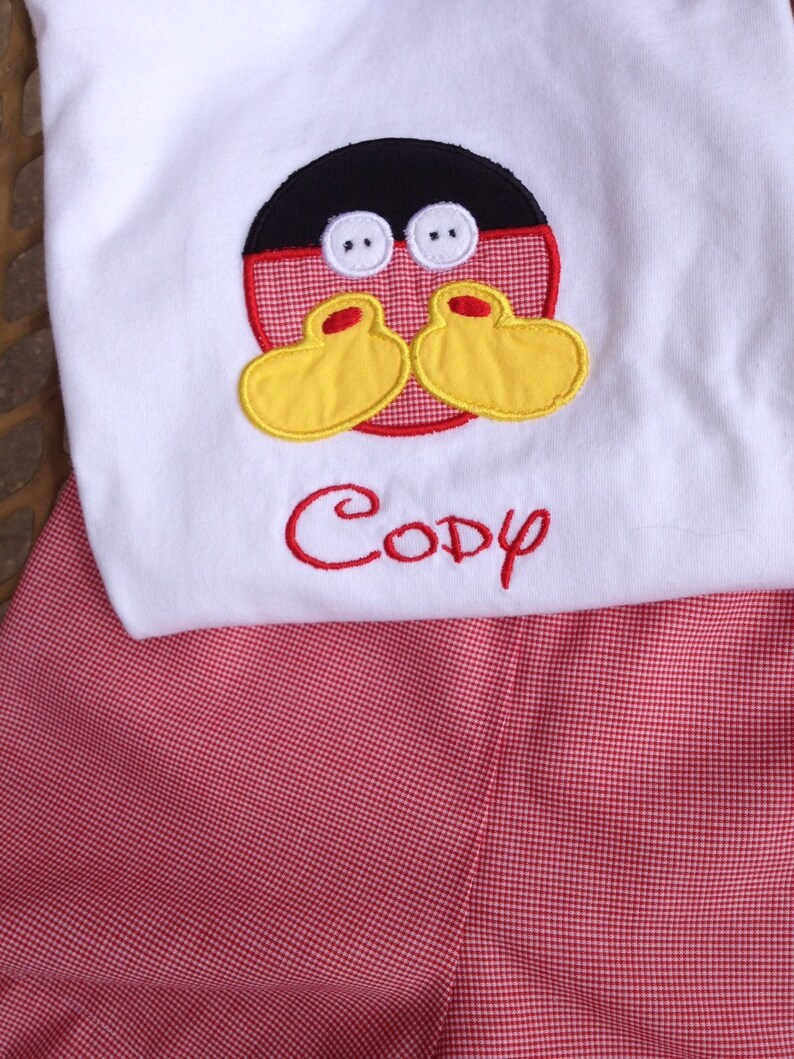 May include: White top with a Mickey Mouse face applique and the name "Cody" embroidered in red. The outfit includes red and white checkered shorts, creating a playful and coordinated look.