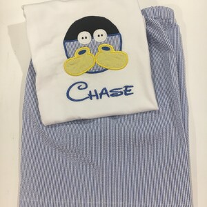 May include: A white pajama top with an embroidered cartoon duck face and the name "CHASE" in blue thread, paired with blue and white striped pajama pants. The duck has a black head, yellow feet, and white eyes.