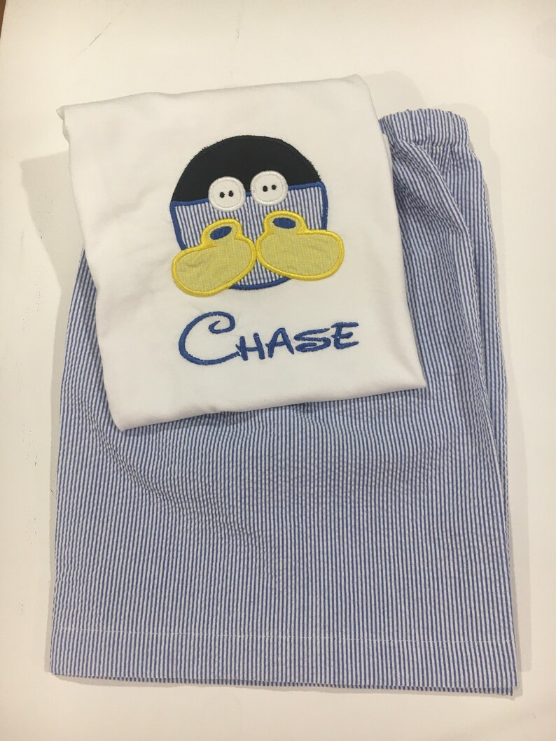 May include: A white pajama top with an embroidered cartoon duck face and the name "CHASE" in blue thread, paired with blue and white striped pajama pants. The duck has a black head, yellow feet, and white eyes.