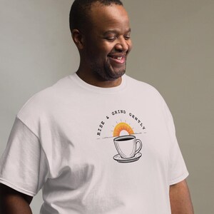 May include: White t-shirt with a coffee cup and sunrise graphic. The text "RISE & GRIND GENTLY" is above the cup. The shirt is a relaxed fit and made of a soft material.