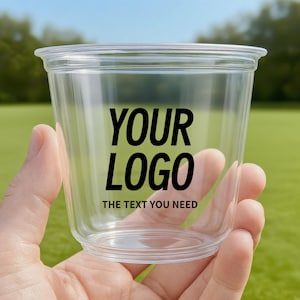 May include: A clear plastic cup with a lid, held in a hand. The cup has the words "YOUR LOGO" in bold black letters, with "THE TEXT YOU NEED" below. The background is a blurred green field and blue sky.